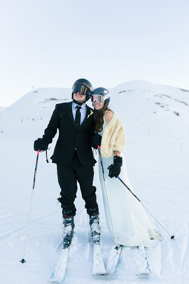 A couple wearing formal wedding attire and ski helmets stand together on skis in a snowy mountain landscape, ready for ski wedding videographer Story Of Your Day to capture their adventurous celebration.
