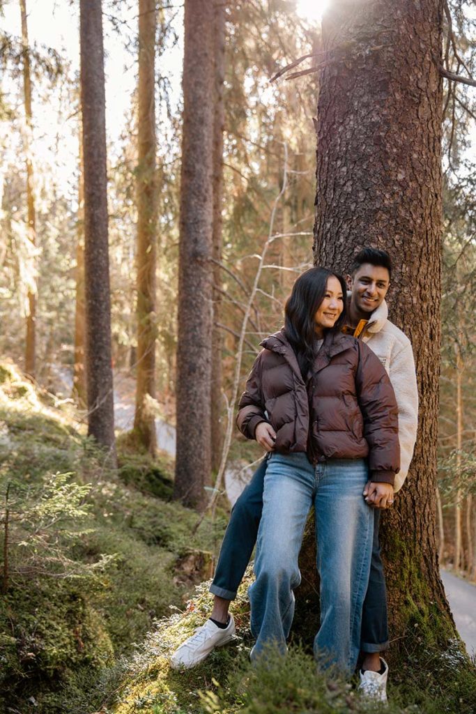 Two people stand close together near a tree in a sunlit forest in Wengen, both wearing jackets and jeans. Surrounded by greenery and tall trees during their proposal shoot