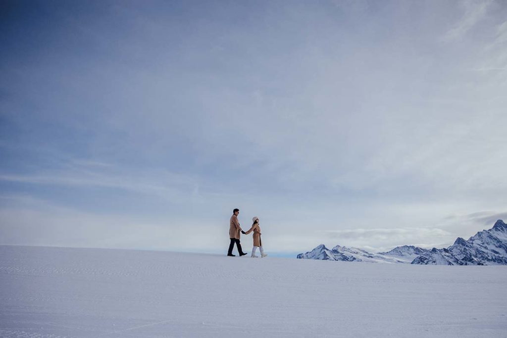 Two people in winter coats walk hand in hand across a snowy landscape on Mannlichen