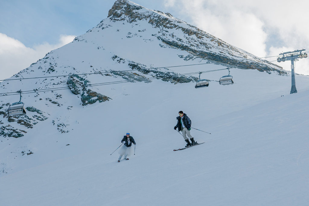 Two people ski down a snowy slope near a ski lift, with a mountain peak and partly cloudy sky in the background.