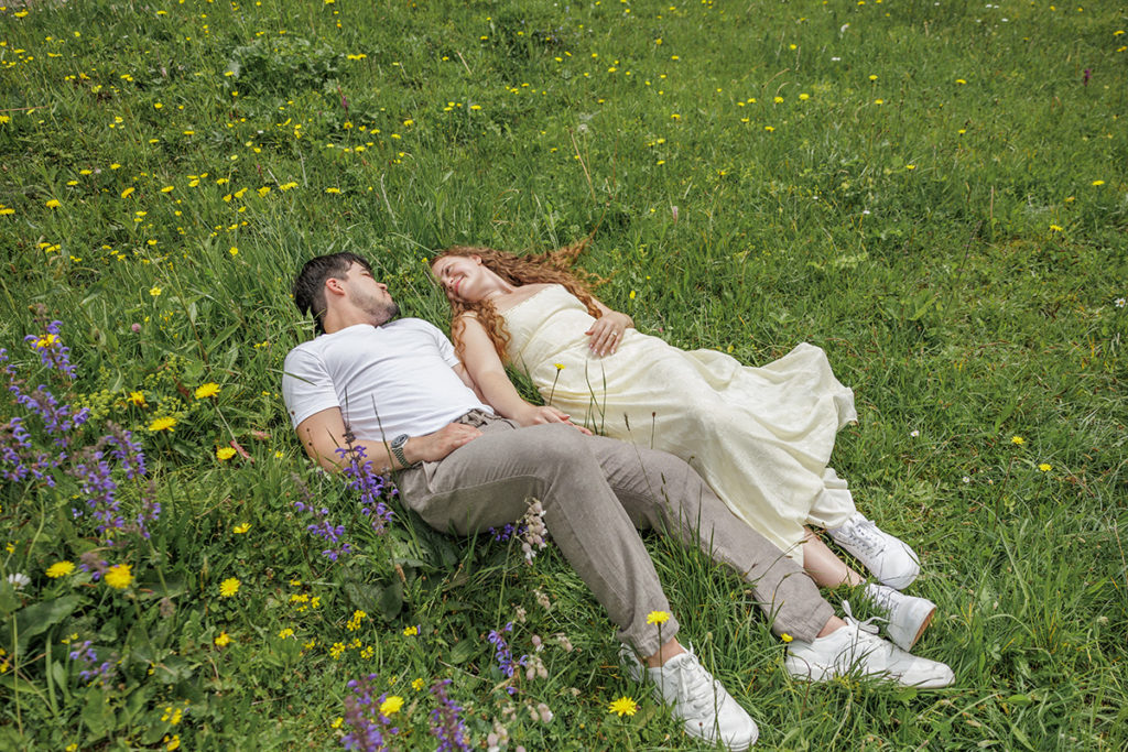 A man and a woman lie on a grassy field in Switzerland with yellow and purple wildflowers, looking at each other and holding hands—a moment like the film Twilight.