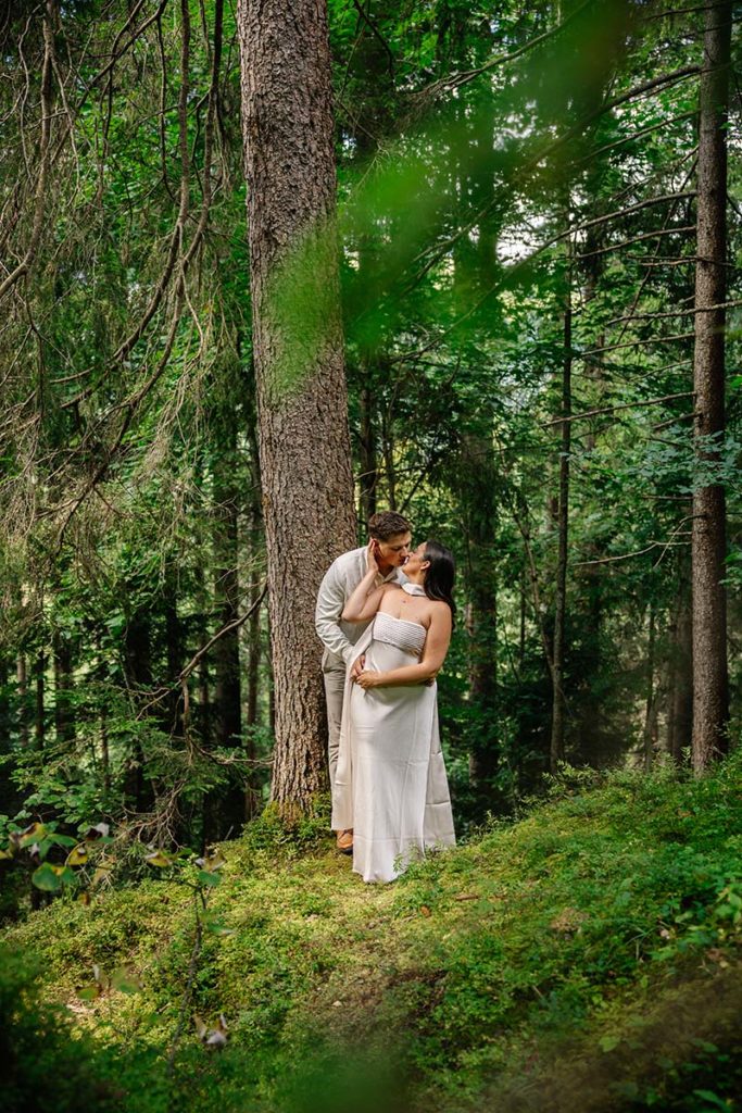 A couple in light clothing stands close together in a green forest, surrounded by trees near Wengen