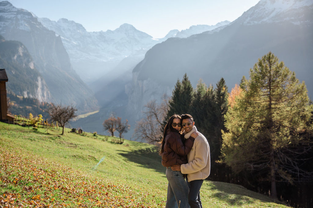 Two people stand closely together on a grassy slope with mountains and trees in the background under a clear sky