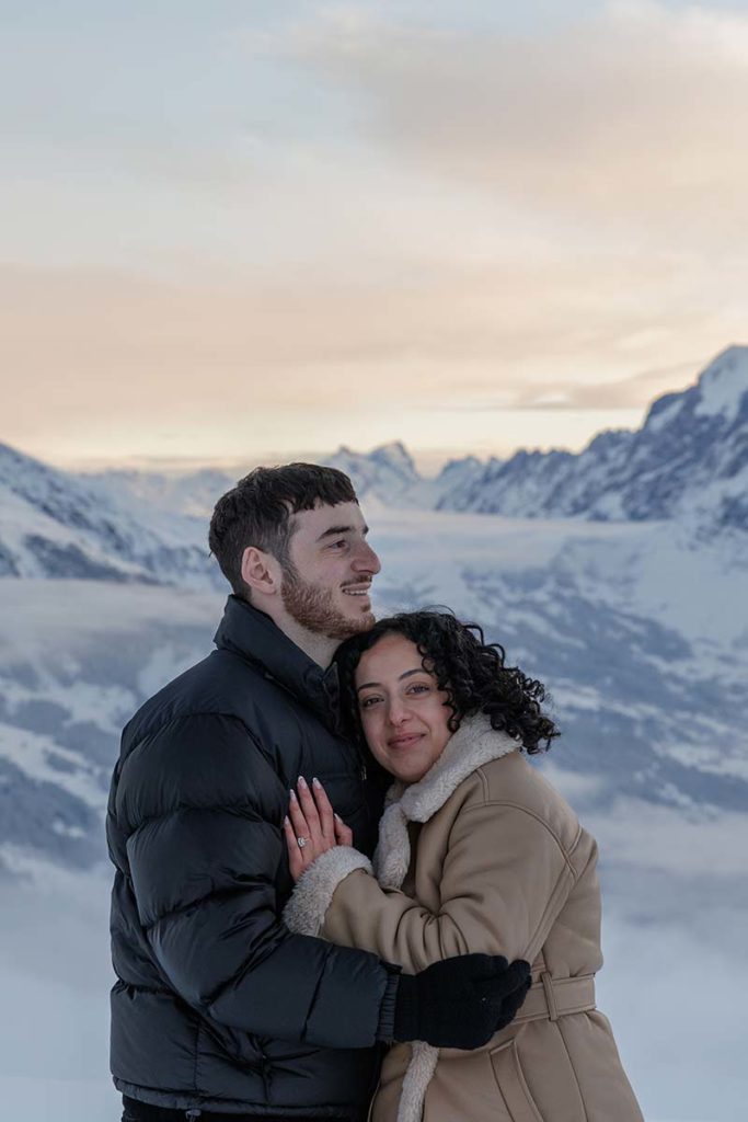 A man and woman in winter coats embrace outdoors, capturing heartfelt engagement moments with snow-covered mountains and a cloudy sky in Lauterbrunnen Valley.