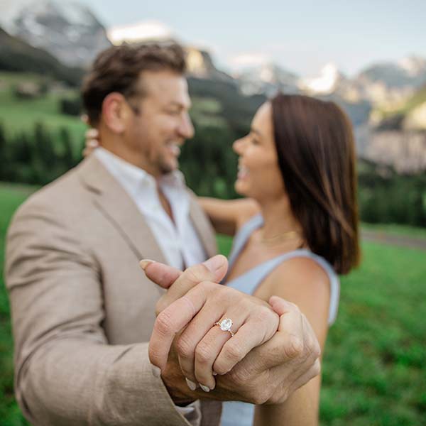 A couple stands outdoors holding hands, the woman showing off her engagement ring. Both are smiling and dressed in semi-formal attire, with mountains and greenery in the background—perfect for stunning destination wedding videography.