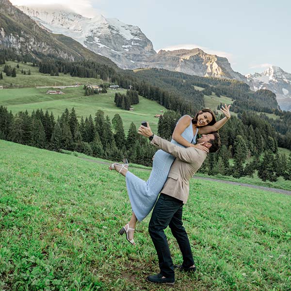 A man lifts a smiling woman in a blue dress in a grassy field, with mountains and trees behind them—a picture-perfect moment reminiscent of destination wedding videography.