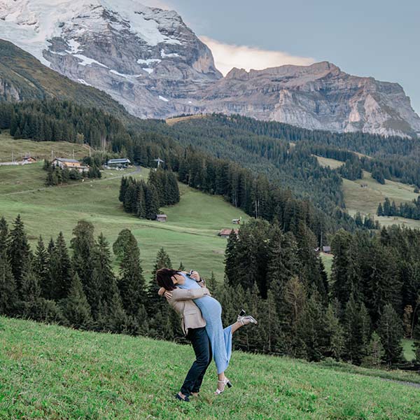 A couple embraces on a grassy hillside with pine trees and snow-capped mountains in the background, making it a perfect setting for destination wedding videography.