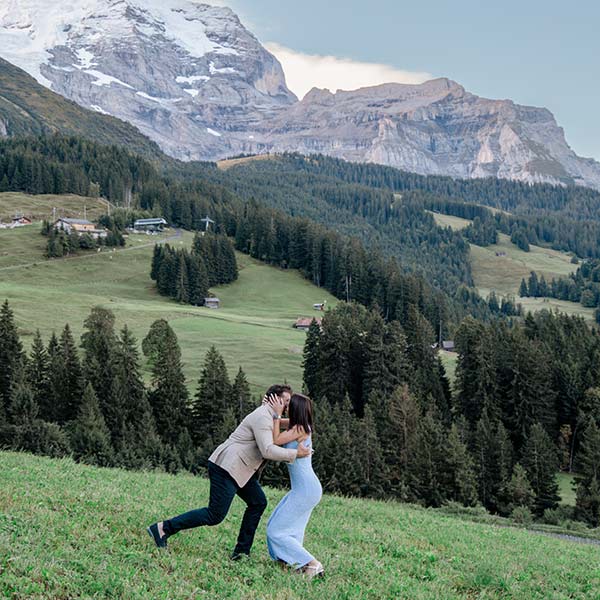A couple stands embracing and laughing in a grassy field with pine trees and snow-capped mountains behind them, capturing the magic of destination wedding videography.