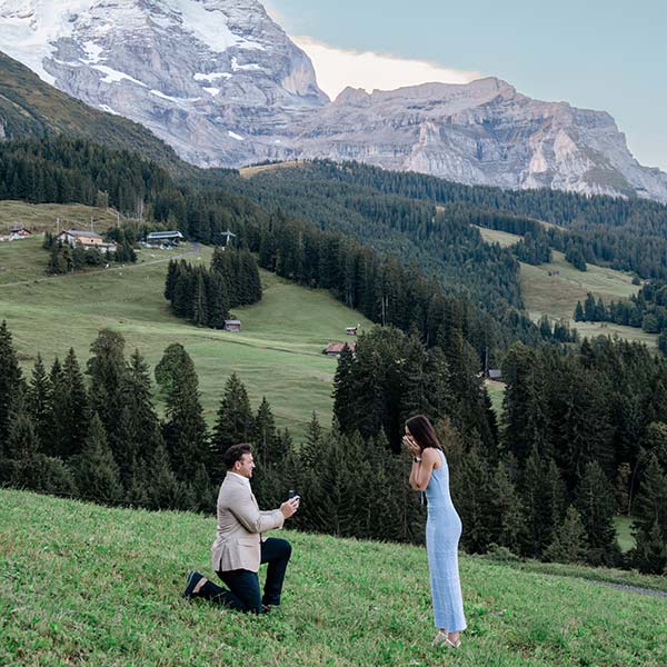 A man kneels and proposes to a woman in a grassy field with mountains and trees in the background, capturing a perfect moment for destination wedding videography.
