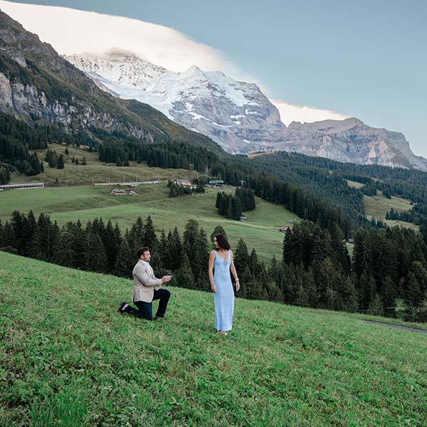 A man kneels on one knee proposing to a woman on a grassy hillside, with mountains and trees in the background—an unforgettable moment perfect for wedding videography or a dream destination wedding.