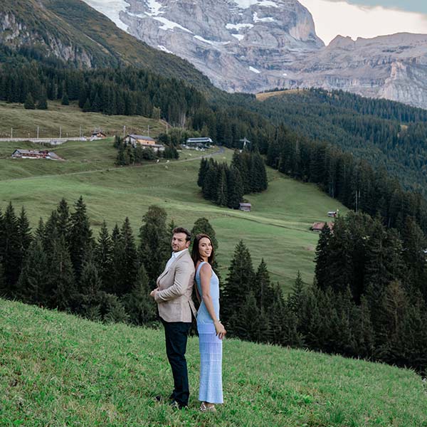 A man and woman stand back to back on a grassy hillside with forest, scattered houses, and mountains in the background—an idyllic scene perfect for destination videography.