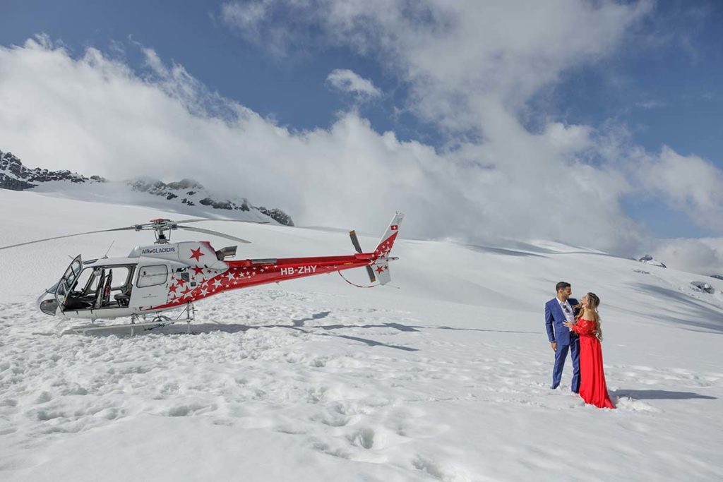 A red and white helicopter is parked on snow as a man in a blue suit stands beside a woman in a red dress, capturing an unforgettable Engagement Lauterbrunnen Valley moment amid snowy mountains and clouds.