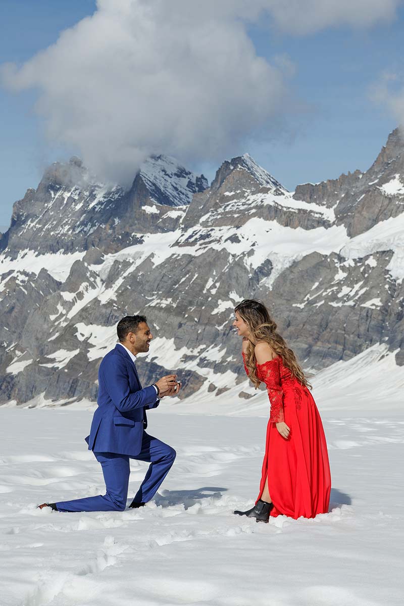 A man in a blue suit kneels on snow proposing to a woman in a red dress, capturing heartwarming proposal moments against the backdrop of snow-covered mountains.