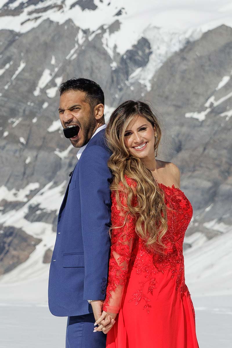 A man in a blue suit and a woman in a red dress stand back to back, holding hands, sharing an Engagement Lauterbrunnen Valley moment with snowy mountains in the background.