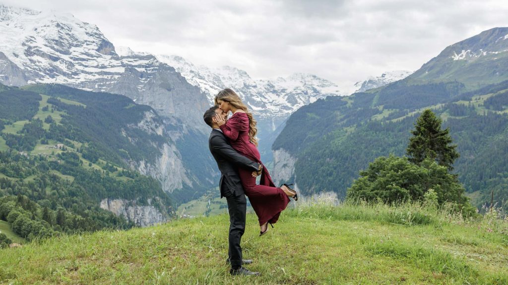 A man in a suit lifts and kisses a woman in a red dress on a grassy hill, celebrating their engagement in Lauterbrunnen Valley, with snow-capped mountains and a stunning valley as the backdrop.