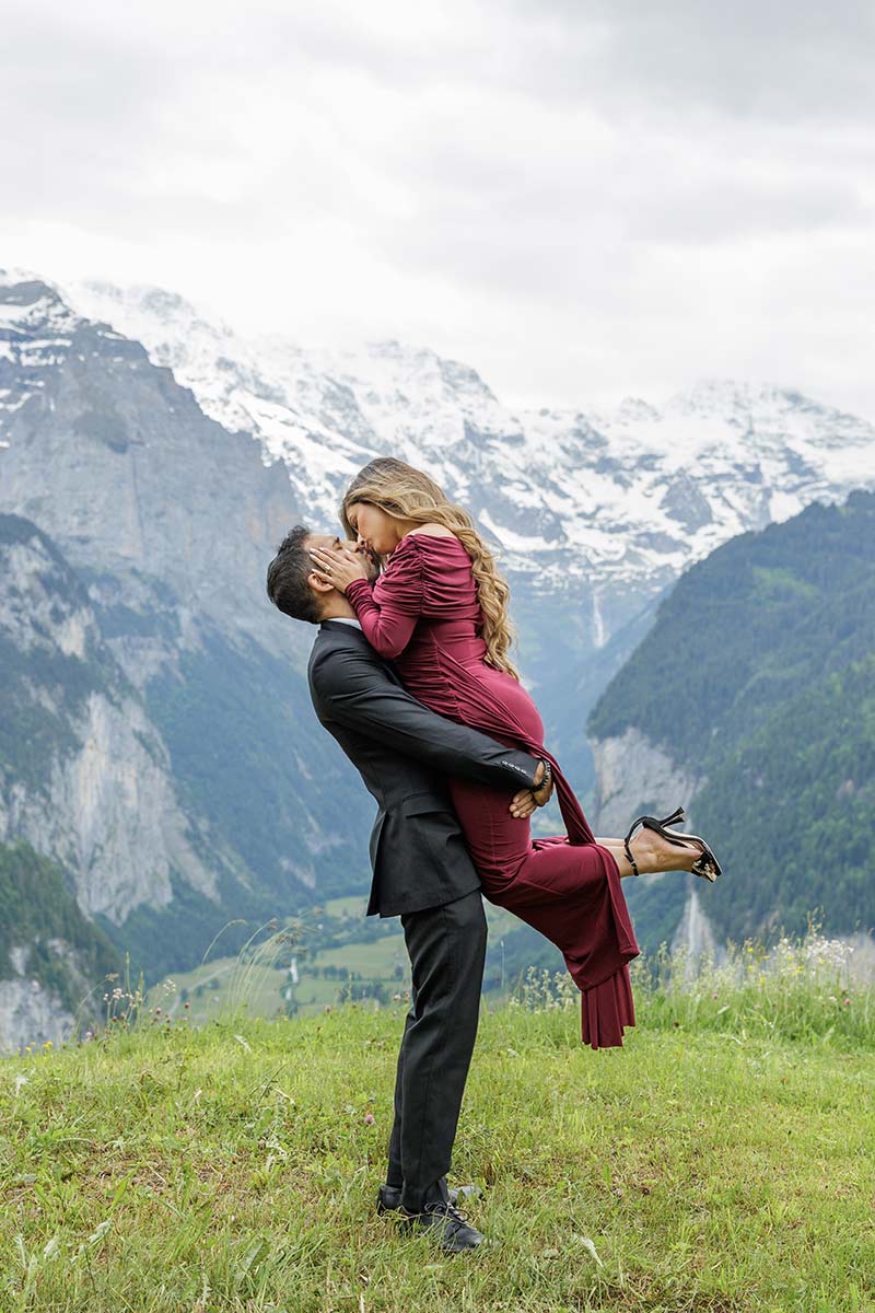 A man in a suit lifts and kisses a woman in a red dress on a grassy hill, capturing unforgettable engagement Wengen moments with snow-capped mountains rising behind them.