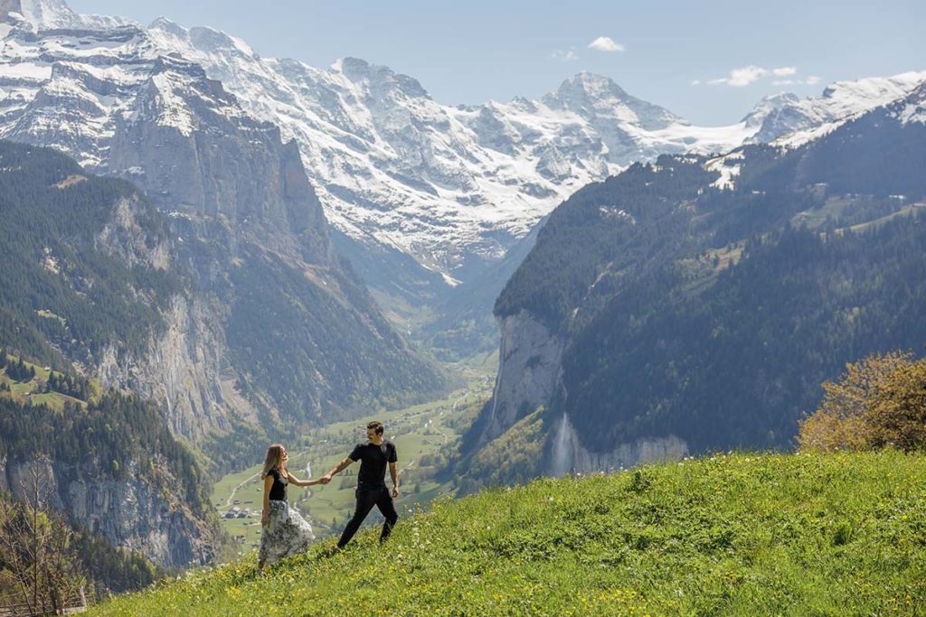A man and woman walk hand in hand on a grassy hill with the stunning Lauterbrunnen Valley and snow-capped mountains as their backdrop, capturing unforgettable engagement moments.