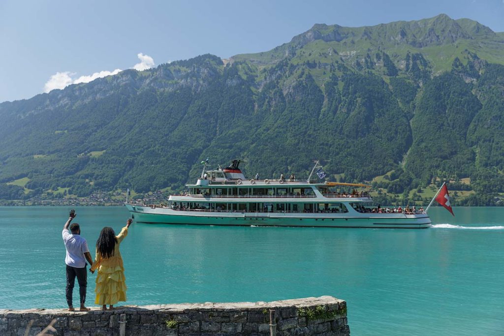 A man and a woman stand by a lakeside in the Lauterbrunnen Valley, waving at a large passenger boat sailing on turquoise water with mountains in the background.