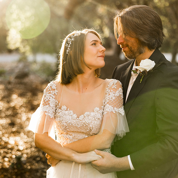 A bride and groom stand outdoors, embracing and looking at each other as sunlight filters through the trees during their celebrity Santa Barbara wedding