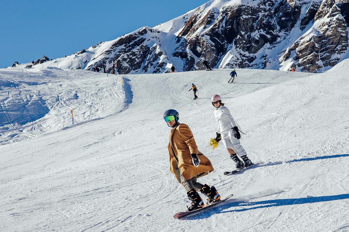 A bride and groom snowboard and ski down a snowy mountain slope in Switzerland under a clear blue sky, with rocky peaks visible in the background during their Swiss adventure ski wedding in Wengen