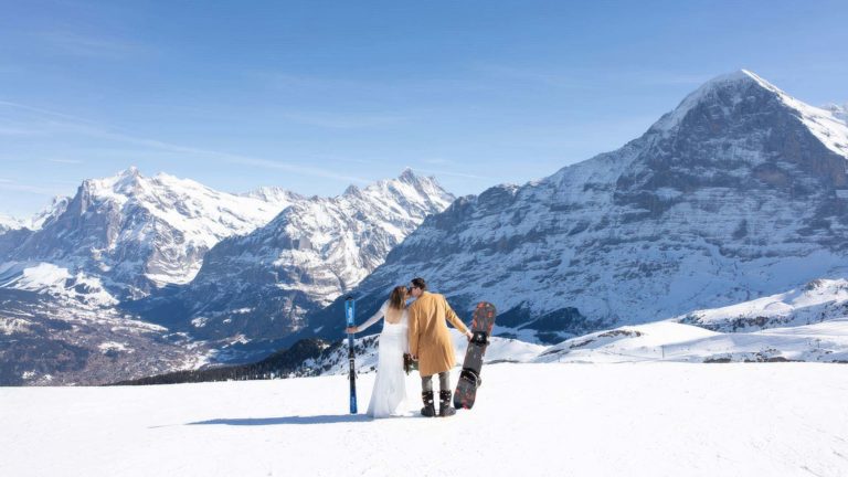 A couple dressed in wedding attire stands on a snowy mountain slope in Switzerland with skis and a snowboard, facing the Eiger under a clear blue sky, during their ski adventure wedding in Wengen in the Swiss Alps