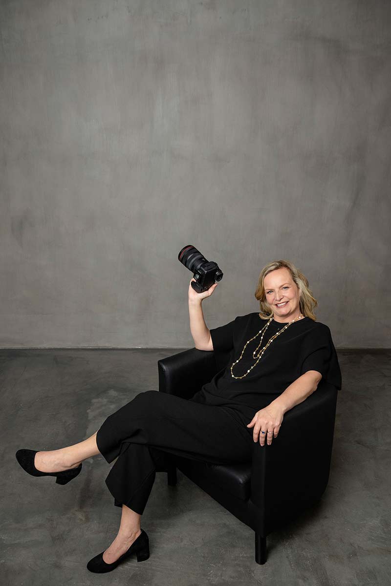 Emma Wilson from Story Of Your Day is seated in a black armchair, smiling and holding a camera up in her right hand against a plain gray background