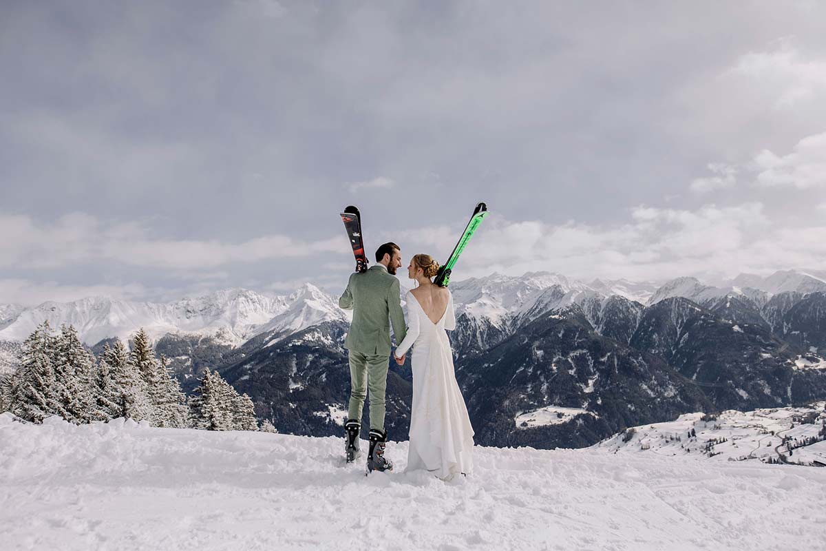 A bride and groom, dressed in wedding attire and holding skis, stand together on a snowy mountain with breathtaking peaks behind them—an unforgettable ski wedding perfect for any adventure wedding film.