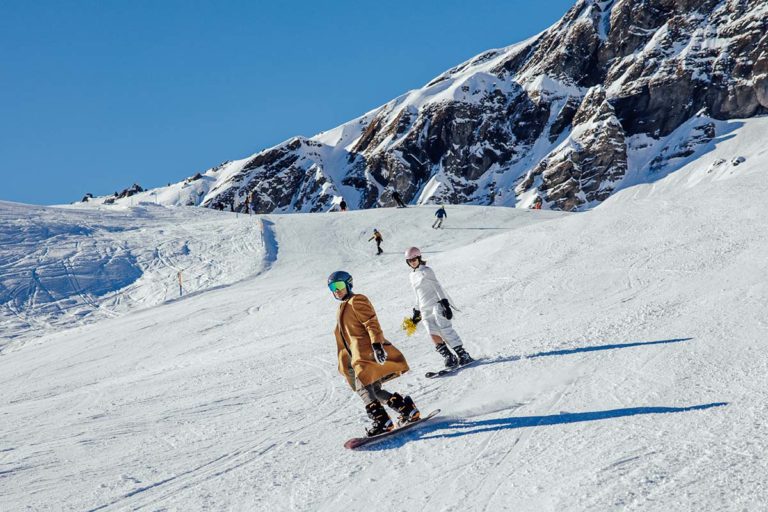 Several snowboarders ride down a snowy mountain slope under a clear blue sky, with rocky, snow-covered peaks in the background—a perfect adventure wedding setting for any ski wedding film portfolio.