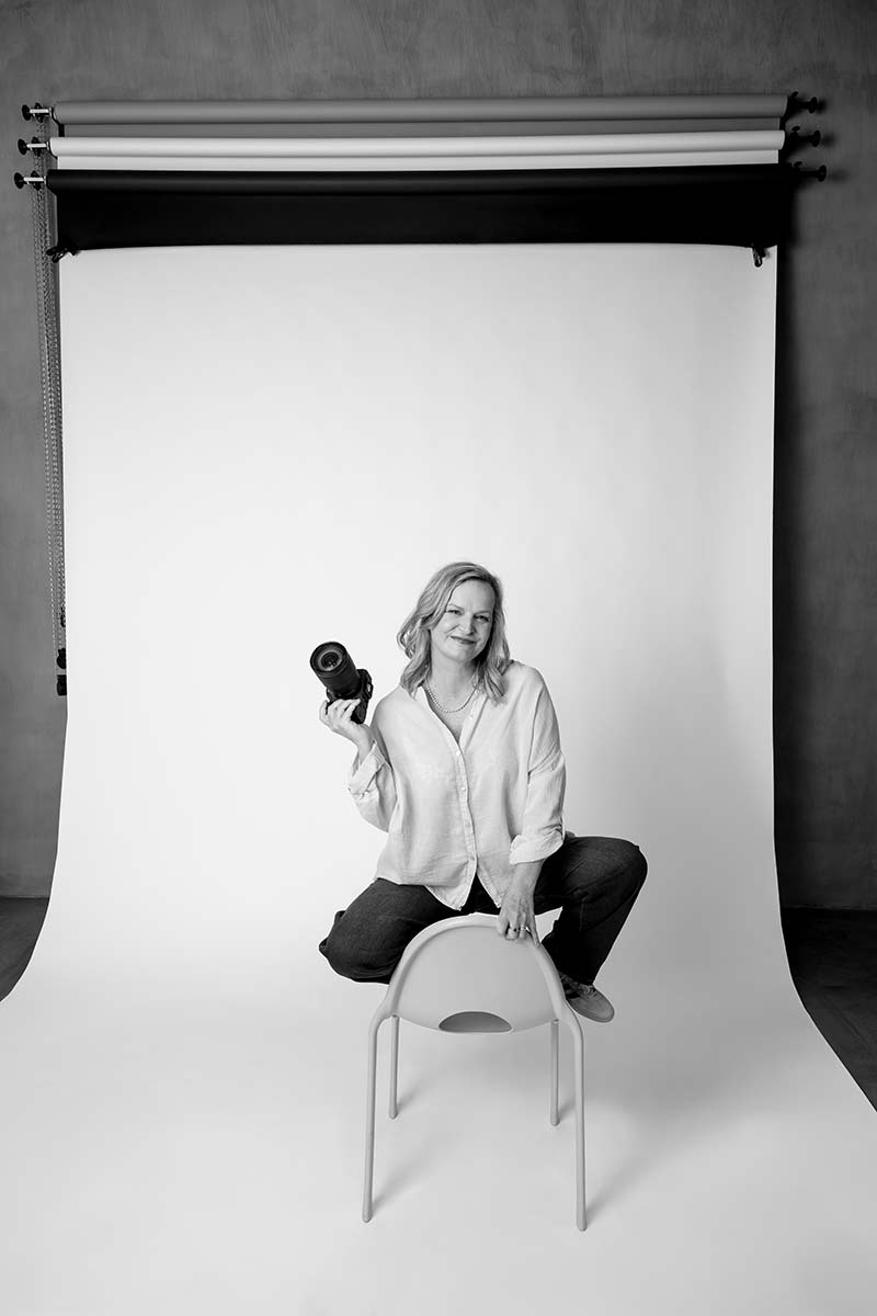 A woman sits on a chair in front of a photography backdrop, holding a camera and smiling. The photo is black and white.