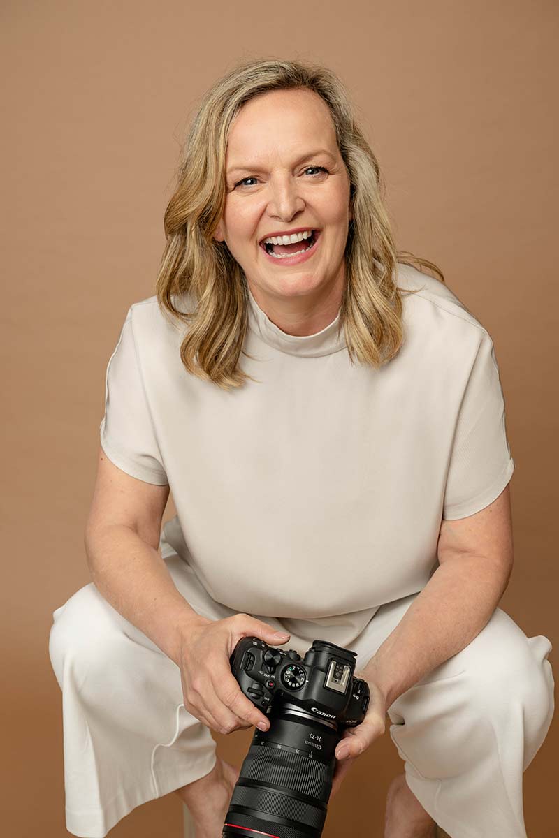A smiling woman with blonde hair in a light outfit holds a professional camera against a plain brown background.