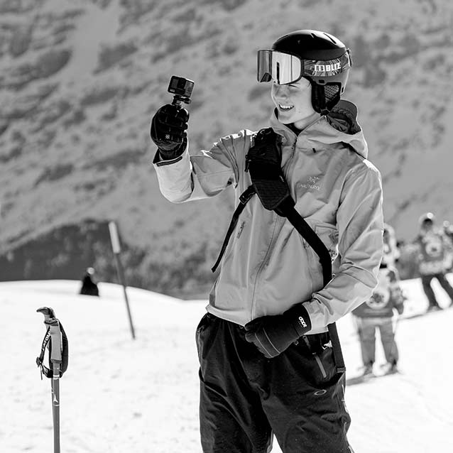 A person wearing ski gear holds a small action camera on a snowy mountain slope, with other skiers and snow-covered peaks in the background.