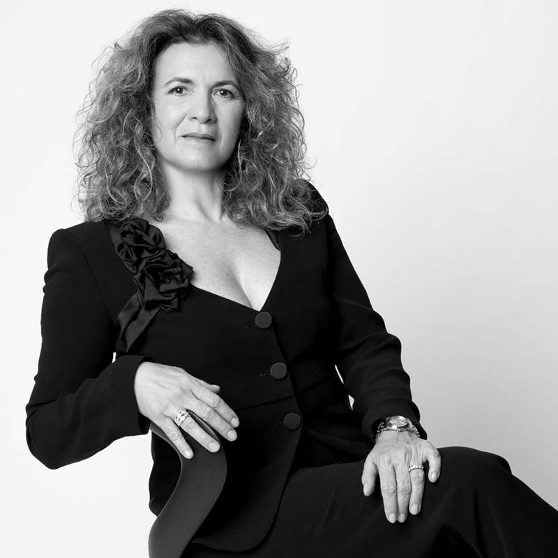 A woman with curly hair wearing a dark suit jacket sits on a chair, looking at the camera with a neutral expression, as if ready to share the story of your day against a plain background.