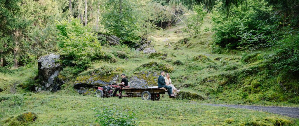 Two people sit on a wooden trailer attached to a small motorized tractor, traveling through a lush, green, mossy forest in Wengen