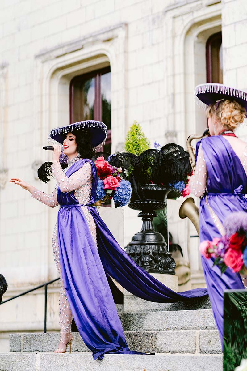 Two women in purple dresses and wide-brimmed hats perform on outdoor steps near a building, surrounded by floral decorations and a microphone.