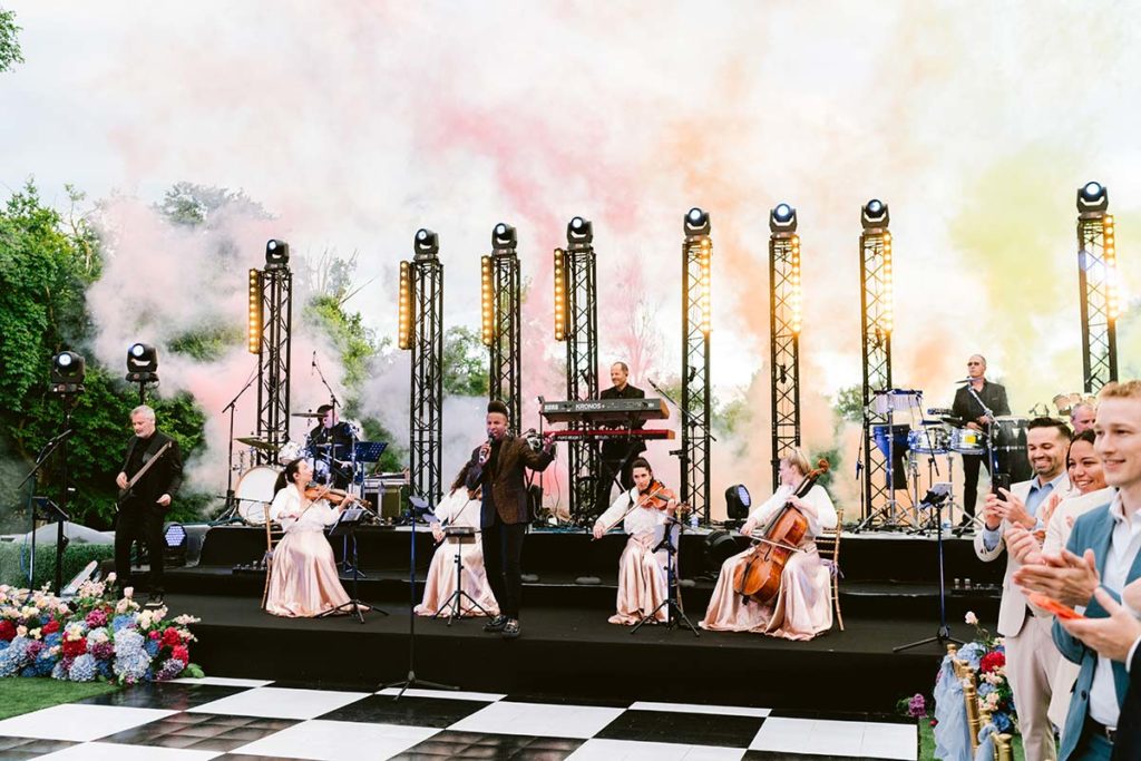 A live band with string and percussion instruments performs on an outdoor stage with colorful smoke and lights, while an applauding audience enjoys the show—perfect for capturing memorable moments through event videography.