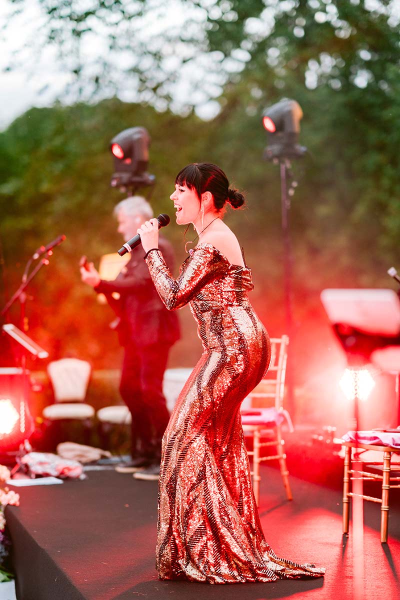 A woman in a shiny, gold, off-the-shoulder dress sings into a microphone on an outdoor stage with red lighting and a guitarist in the background, all captured by expert event videography.