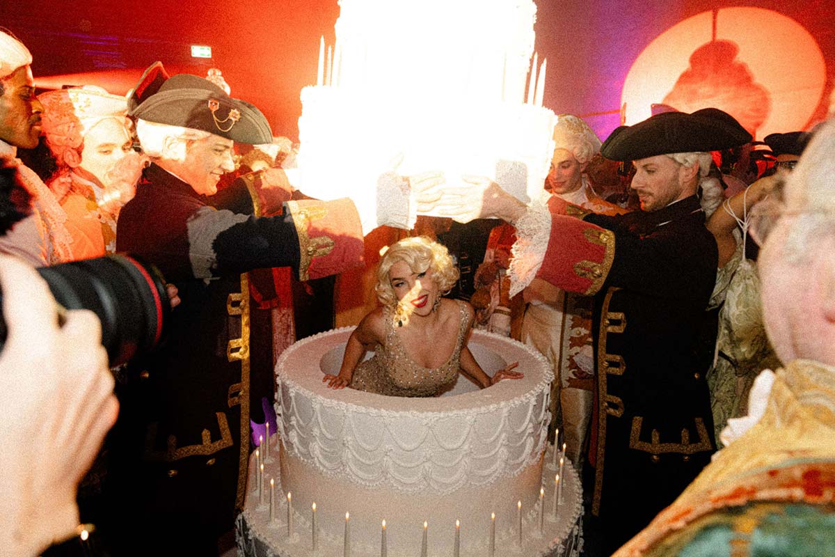 A woman in a sparkling dress emerges from a large decorative cake at a party, surrounded by people in 18th-century-style costumes—an unforgettable scene perfect for event videography.