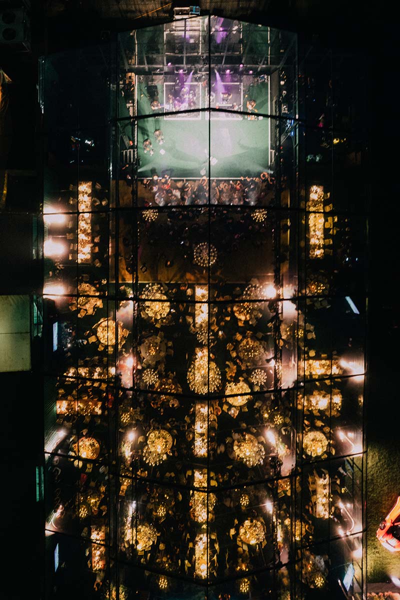 Aerial view of a glass-walled venue with tables arranged for an event, illuminated by warm lighting and filled with seated guests—perfect for capturing stunning Event Videography.