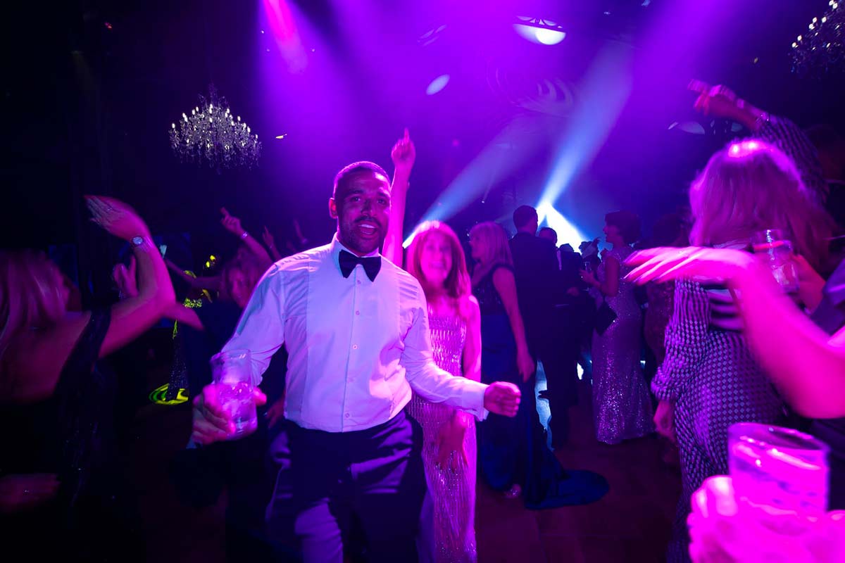 People dancing on a dimly lit dance floor under purple lights at a formal event, captured in perfect detail through expert event videography, with one man in a white shirt and bow tie in the foreground holding a drink.