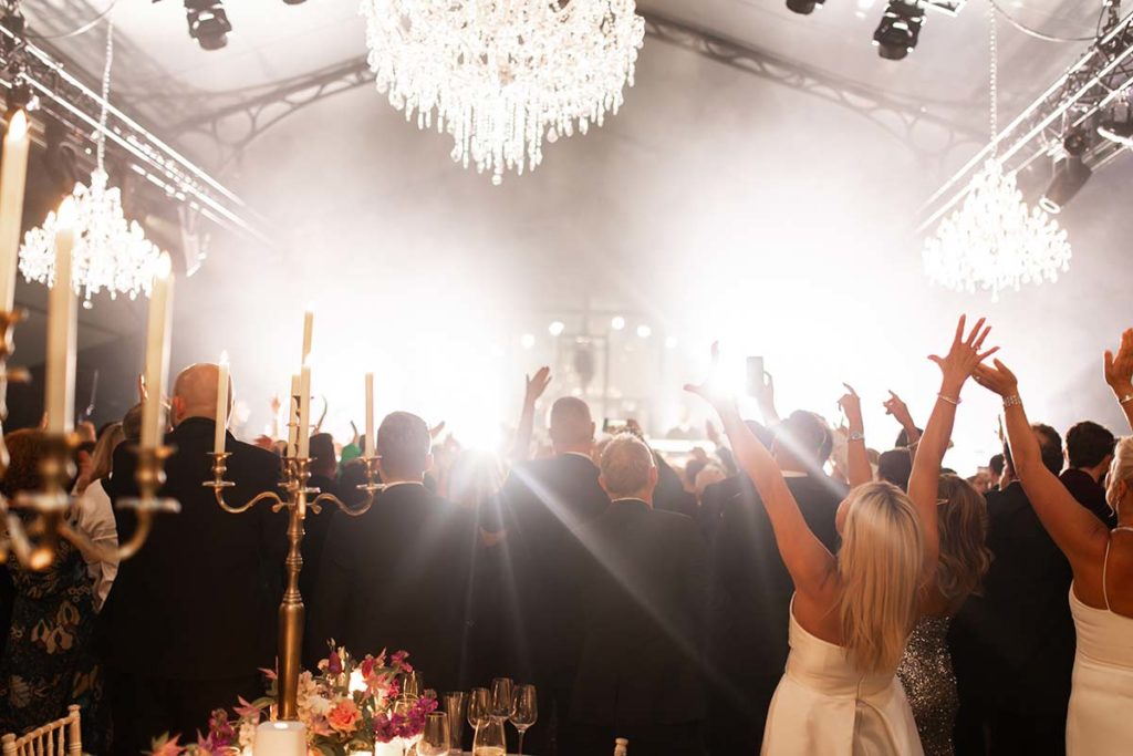 Crowd of people in formal attire with raised hands facing a brightly lit stage under chandeliers at an indoor event venue, captured through expert event videography.