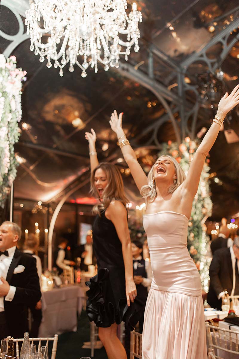 Two women in formal dresses stand on chairs with arms raised, celebrating at an elegant indoor event with chandeliers and decorated tables—perfect moments for event videography to capture the joyful atmosphere.