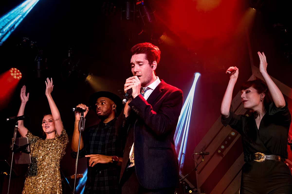 Four musicians perform on stage under colorful lights; the central singer holds a microphone while the others sing and raise their arms, all captured with dynamic event videography.