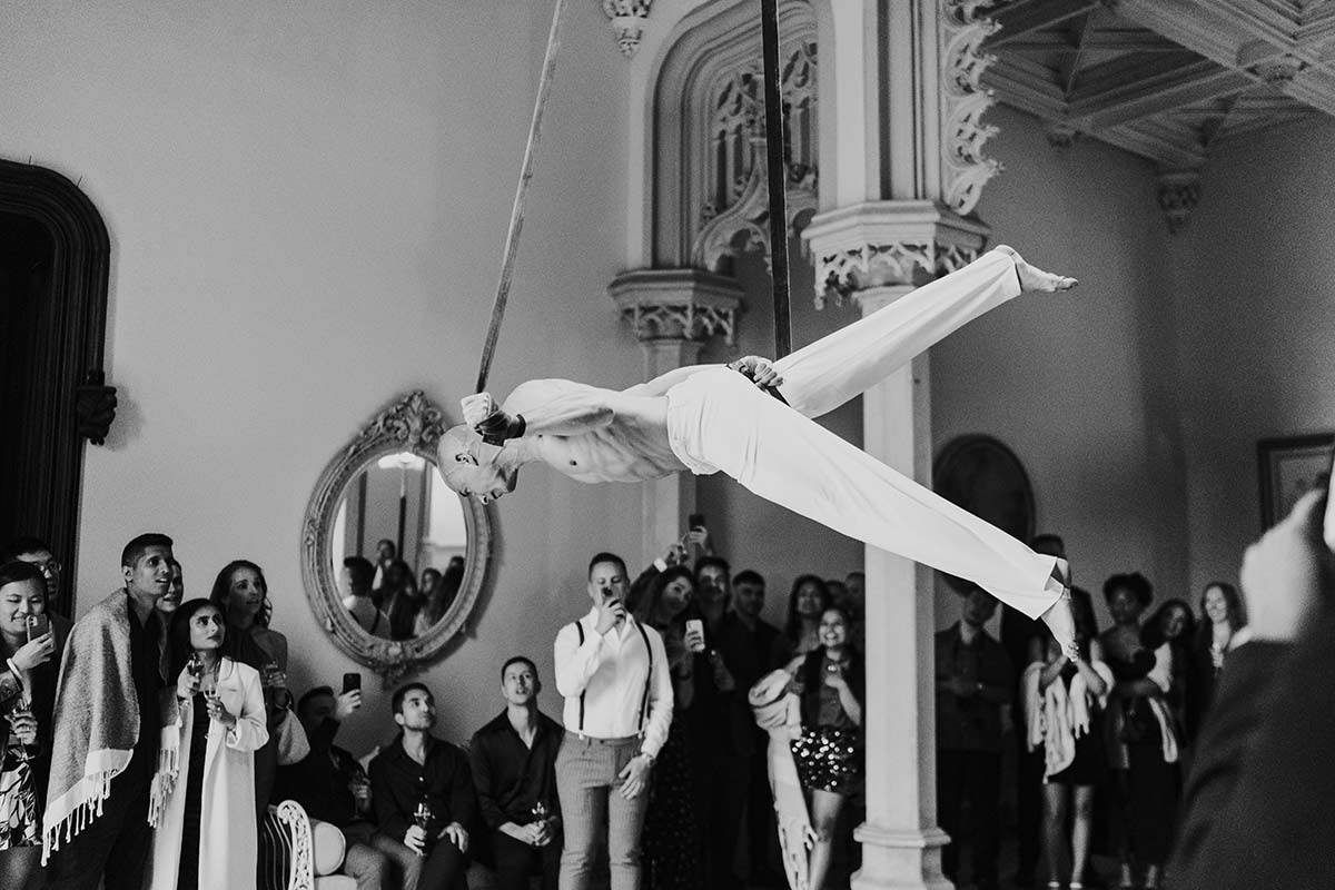 A performer in white clothing is suspended horizontally on aerial silks, captured through event videography, as an audience watches in an ornate room.