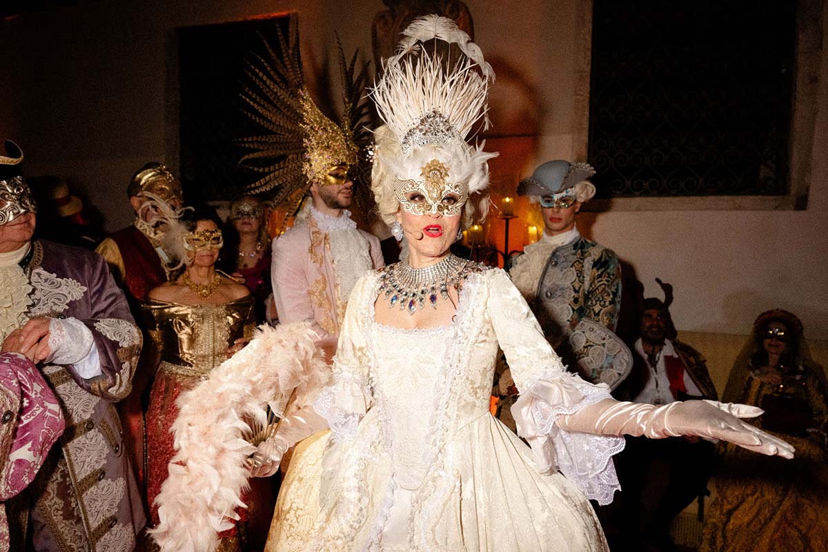 A group of people in ornate, historical costumes and decorative masks attend a lavish masquerade ball indoors, perfectly captured through skilled event videography.
