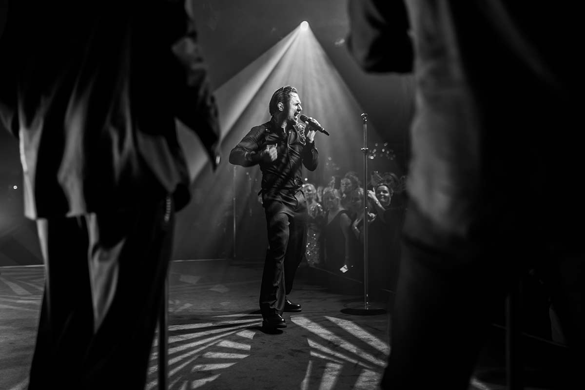 A male performer sings on stage under dramatic lighting, as event videography captures the moment with a crowd and two silhouetted figures visible in the foreground.