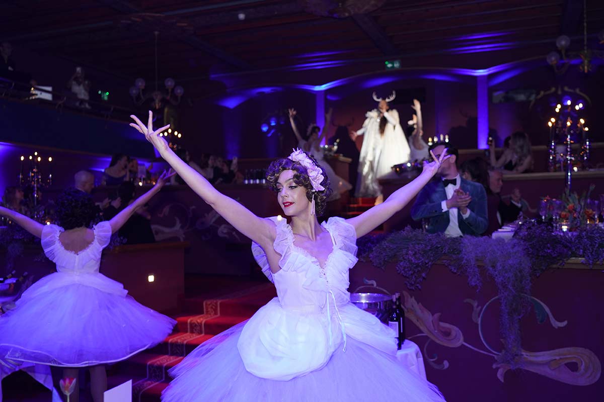 Ballet dancers in white costumes perform gracefully in a dimly lit, elegant venue with purple lighting and an audience at decorated tables—perfect for capturing stunning event videography.