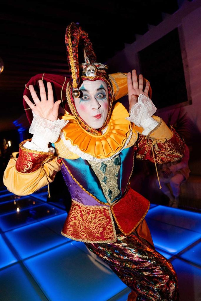Person in a colorful jester costume with white face paint and exaggerated makeup raises both hands near their face, standing on an illuminated floor—perfect for capturing the lively atmosphere in event videography.