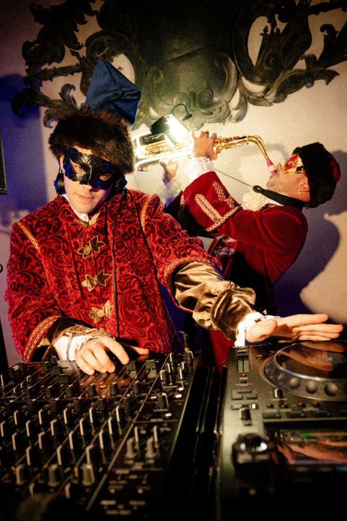 Two people in ornate costumes and masks perform; one operates a DJ mixer in the foreground while the other plays a saxophone in the background, creating an electrifying scene perfect for event videography.