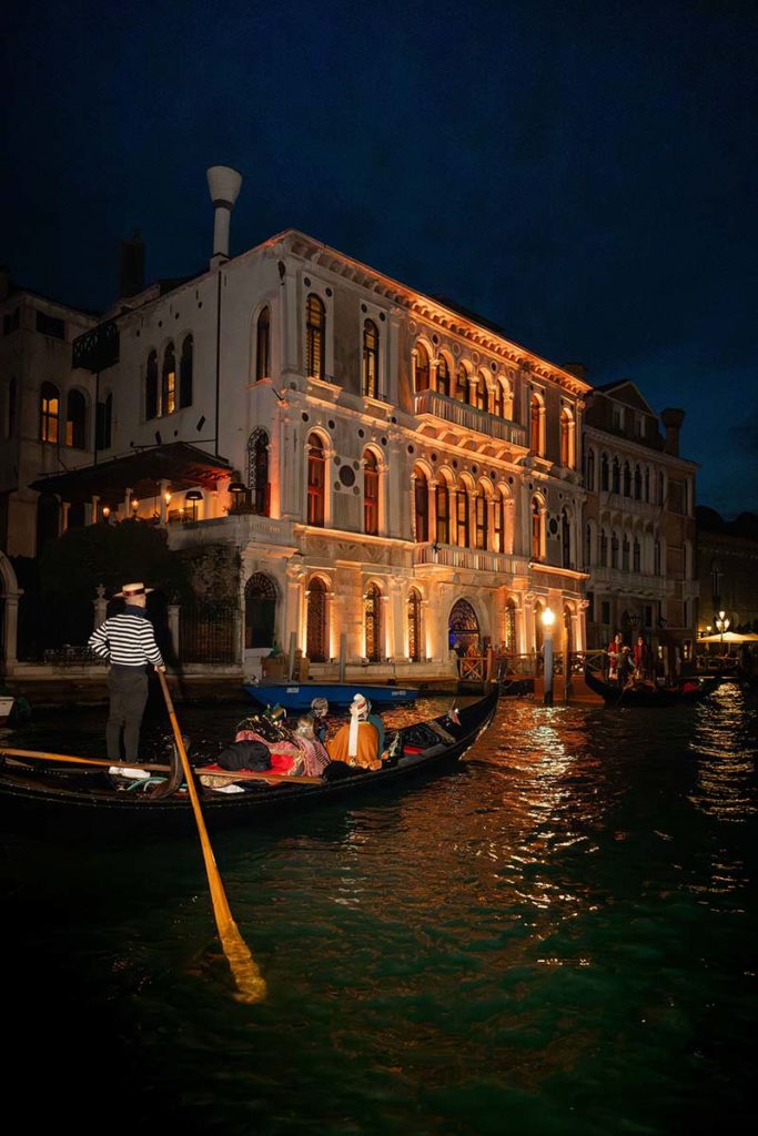 A gondolier steers a gondola carrying passengers along a canal at night, while an event videography crew captures the scene with a brightly lit historic building in the background.