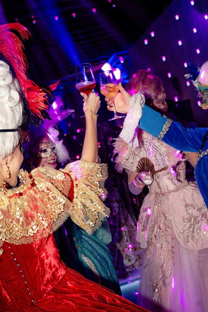 People in elaborate, historical costumes and masks raise glasses in a toast at a lively, colorful masquerade party—perfect for capturing with expert event videography.